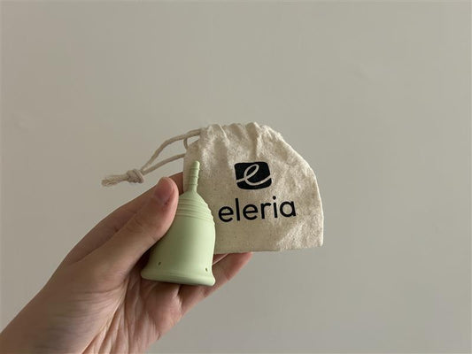 Hand holding a green menstrual cup and cotton drawstring bag. 