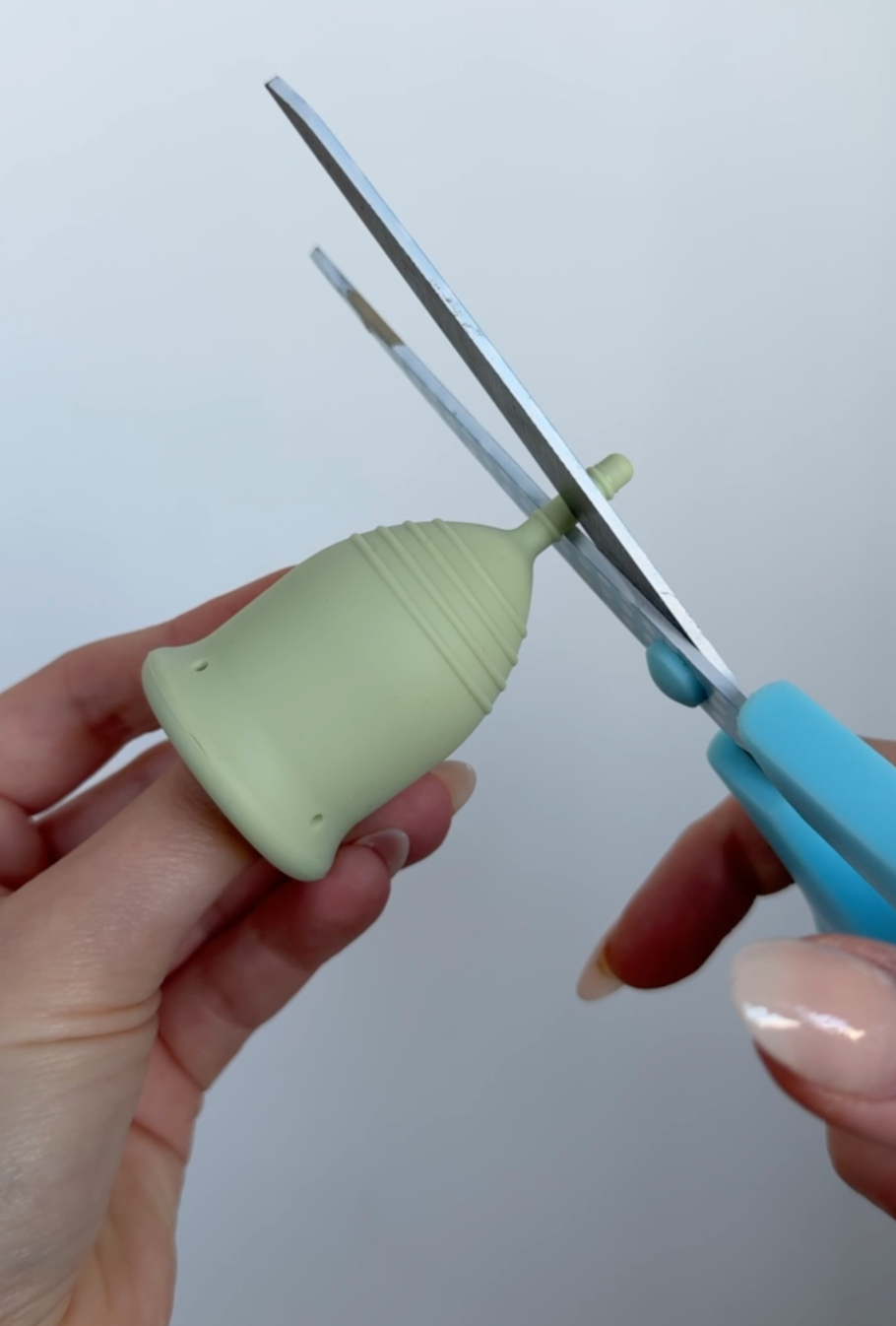 Someone using blue scissors to trim a green period cup stem. 